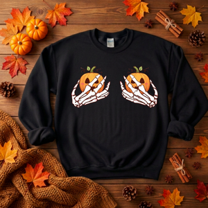 A black crewneck sweatshirt featuring a graphic of two skeleton hands holding orange jack-o'-lantern pumpkins, displayed in a flat-lay autumn setting with fall leaves, pinecones, cinnamon sticks, and a knitted blanket on a wooden surface.