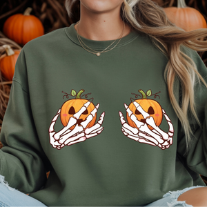 A person wearing an olive green sweatshirt with a Halloween graphic of skeleton hands holding orange jack-o'-lantern pumpkins. The background is filled with pumpkins.
