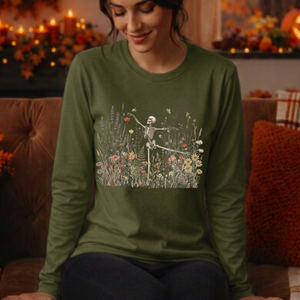 A woman wearing an olive green long-sleeve t-shirt with a graphic of a dancing skeleton in a field of flowers, sitting on a brown couch in a cozy, warmly lit living room.