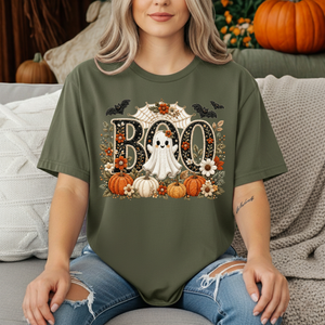 A woman wearing an olive green graphic t-shirt with a Halloween 'BOO' design featuring pumpkins, a ghost, bats, and flowers, sitting on a couch with pillows and pumpkins in the background.
