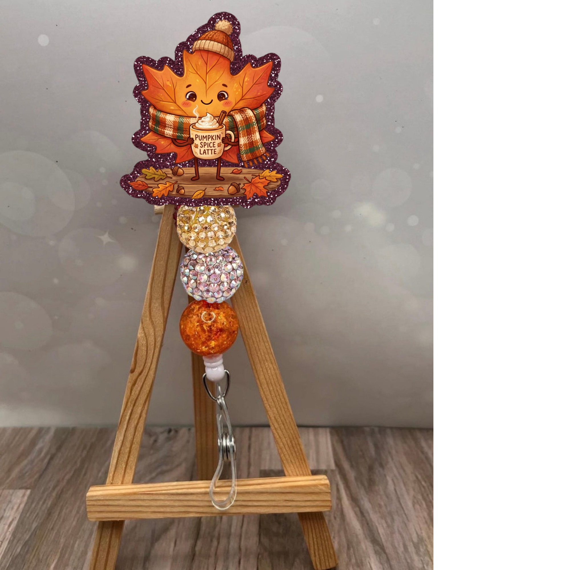 Fall Leaf Badge Pull featuring a cute leaf character with pumpkin spice latte design and glittery beads on a wooden easel.