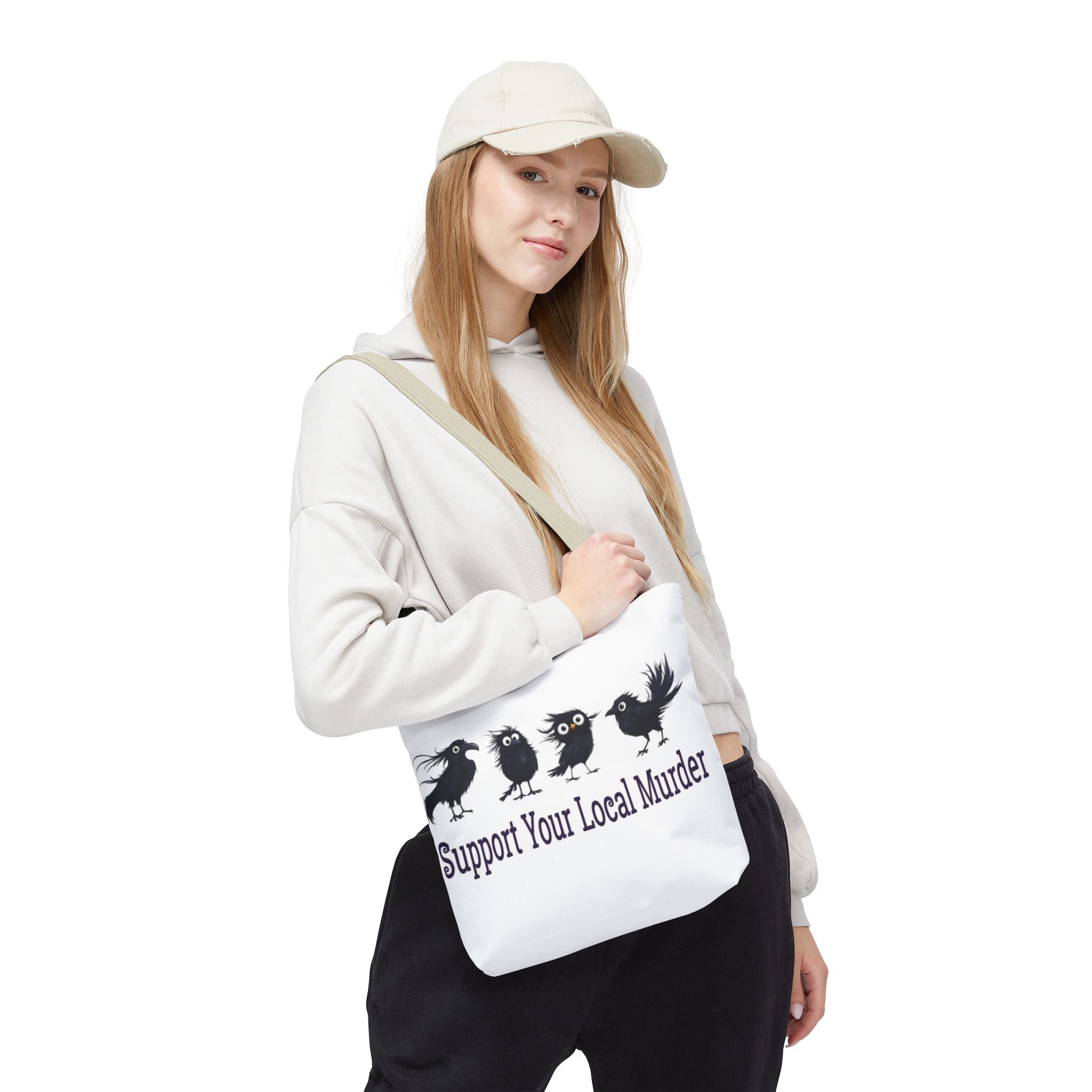A woman wearing a beige cap, white hoodie, and black pants models a white canvas shoulder bag with a black graphic of four crows and the text 'Support Your Local Murder'.