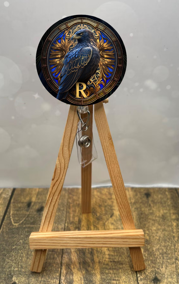 Eagle House badge pull with shimmer glitter featuring a blue raven design displayed on a wooden easel.