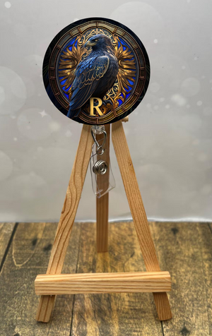 Eagle House badge pull with shimmer glitter featuring a blue raven design displayed on a wooden easel.