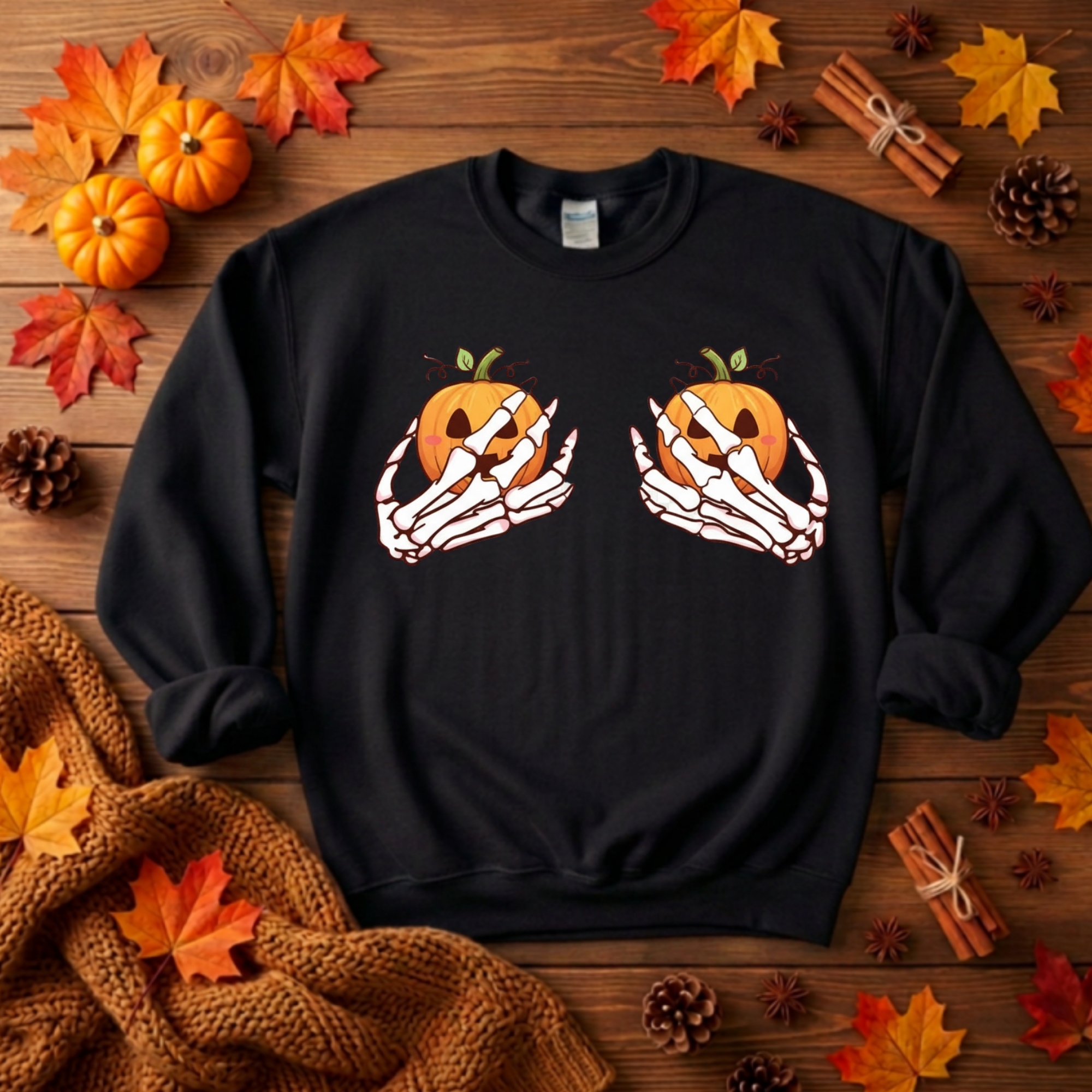 A black crewneck sweatshirt featuring a graphic of two skeleton hands holding orange jack-o'-lantern pumpkins, displayed in a flat-lay autumn setting with fall leaves, pinecones, cinnamon sticks, and a knitted blanket on a wooden surface.