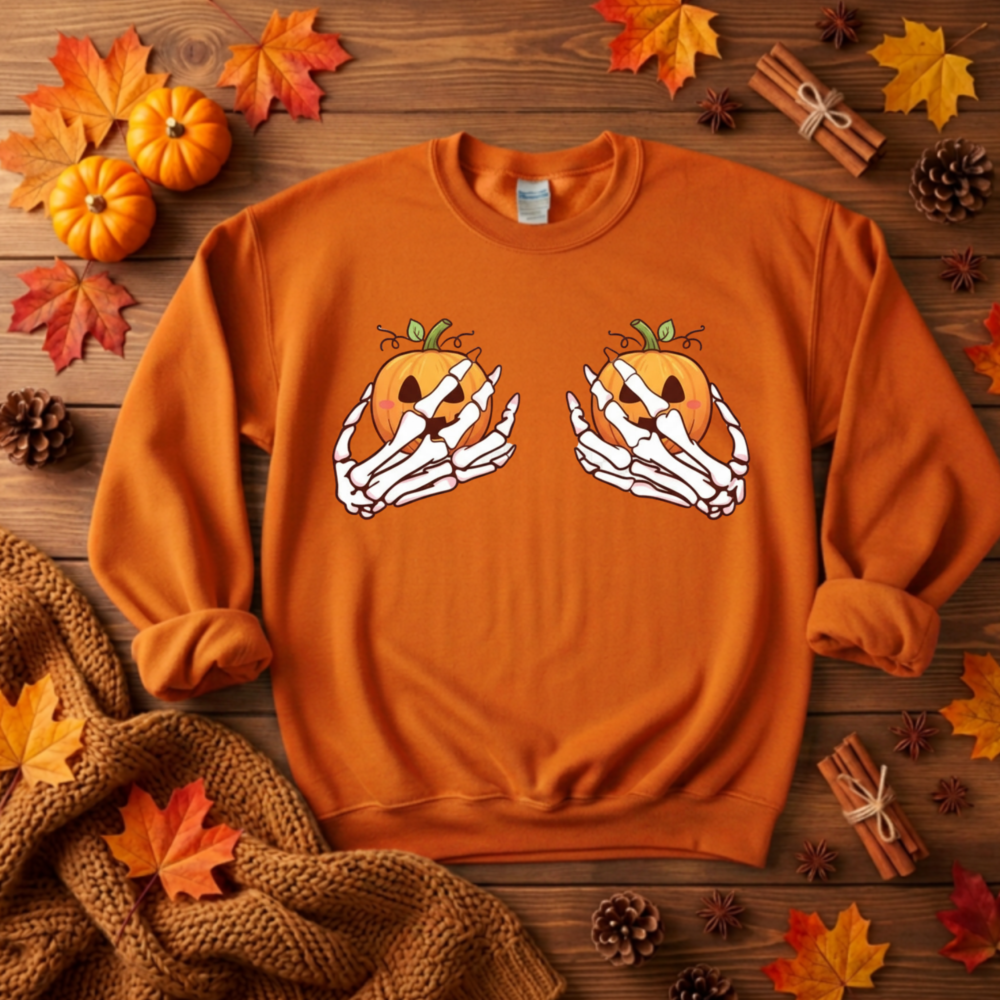 An orange crewneck sweatshirt laid flat on a rustic wooden surface, decorated with autumn leaves, pumpkins, pinecones, cinnamon sticks, and a knitted scarf. The sweatshirt features a graphic of two skeleton hands holding carved jack-o'-lantern pumpkins.