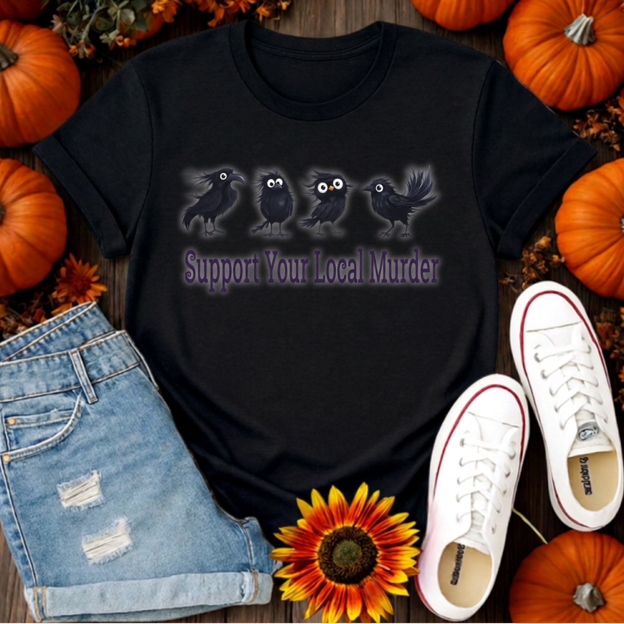A black graphic t-shirt featuring three black birds with glowing eyes and the text 'Support Your Local Murder' in purple. The shirt is styled in a flat-lay arrangement with light-wash denim shorts, white sneakers, a sunflower, and pumpkins on a dark wood background.
