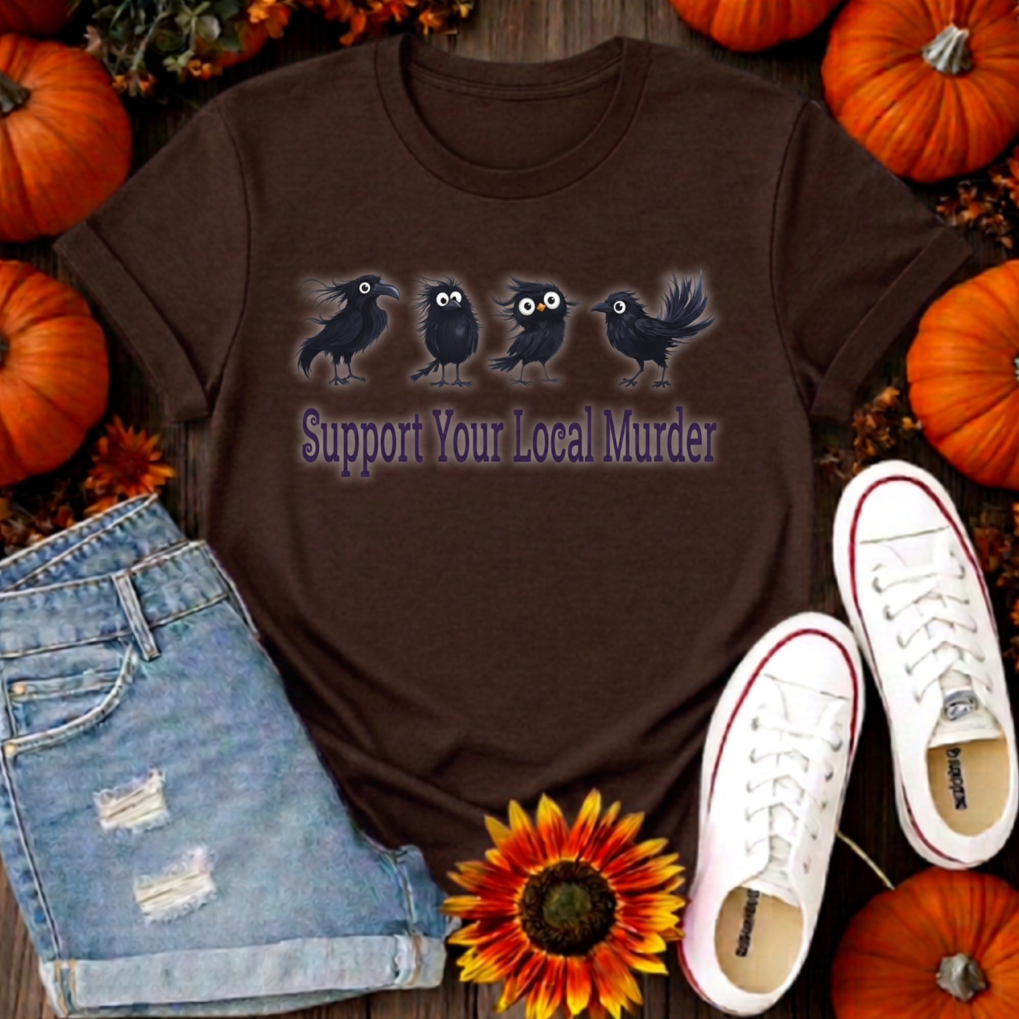 A brown graphic t-shirt featuring four black crows and the text 'Support Your Local Murder' in purple, styled in a flat-lay with denim shorts, white sneakers, pumpkins, and a sunflower.