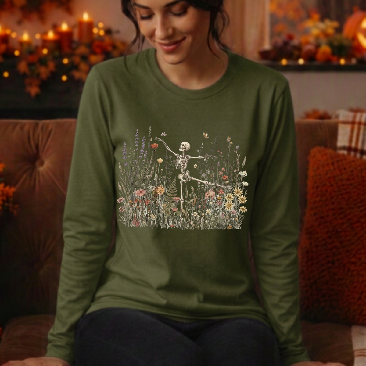 A woman wearing an olive green long-sleeve t-shirt with a graphic of a dancing skeleton in a field of flowers, sitting on a brown couch in a cozy, warmly lit living room.