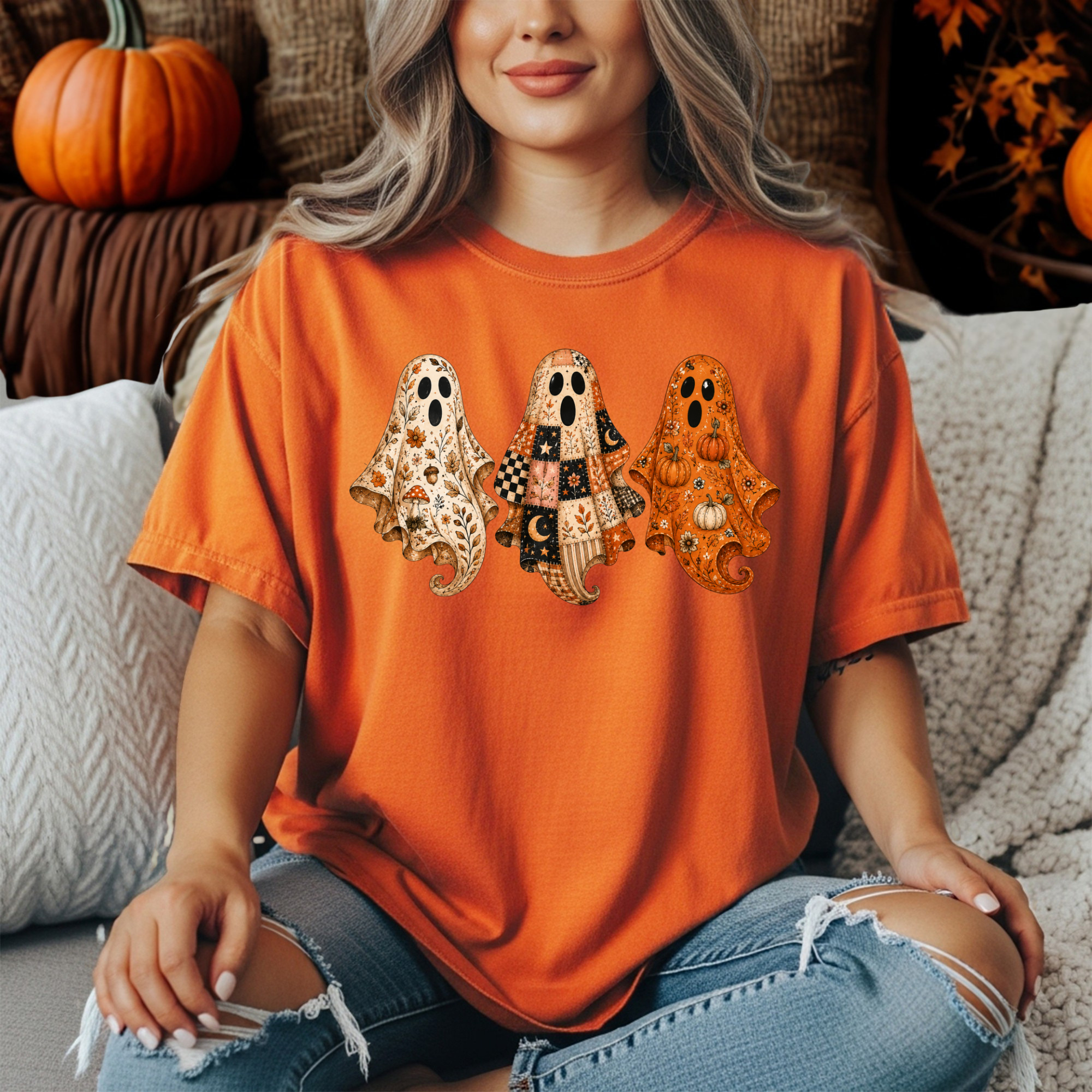 A woman wearing an orange short-sleeve t-shirt with a graphic of three patterned ghosts, sitting on a cozy knit couch with pumpkins in the background.