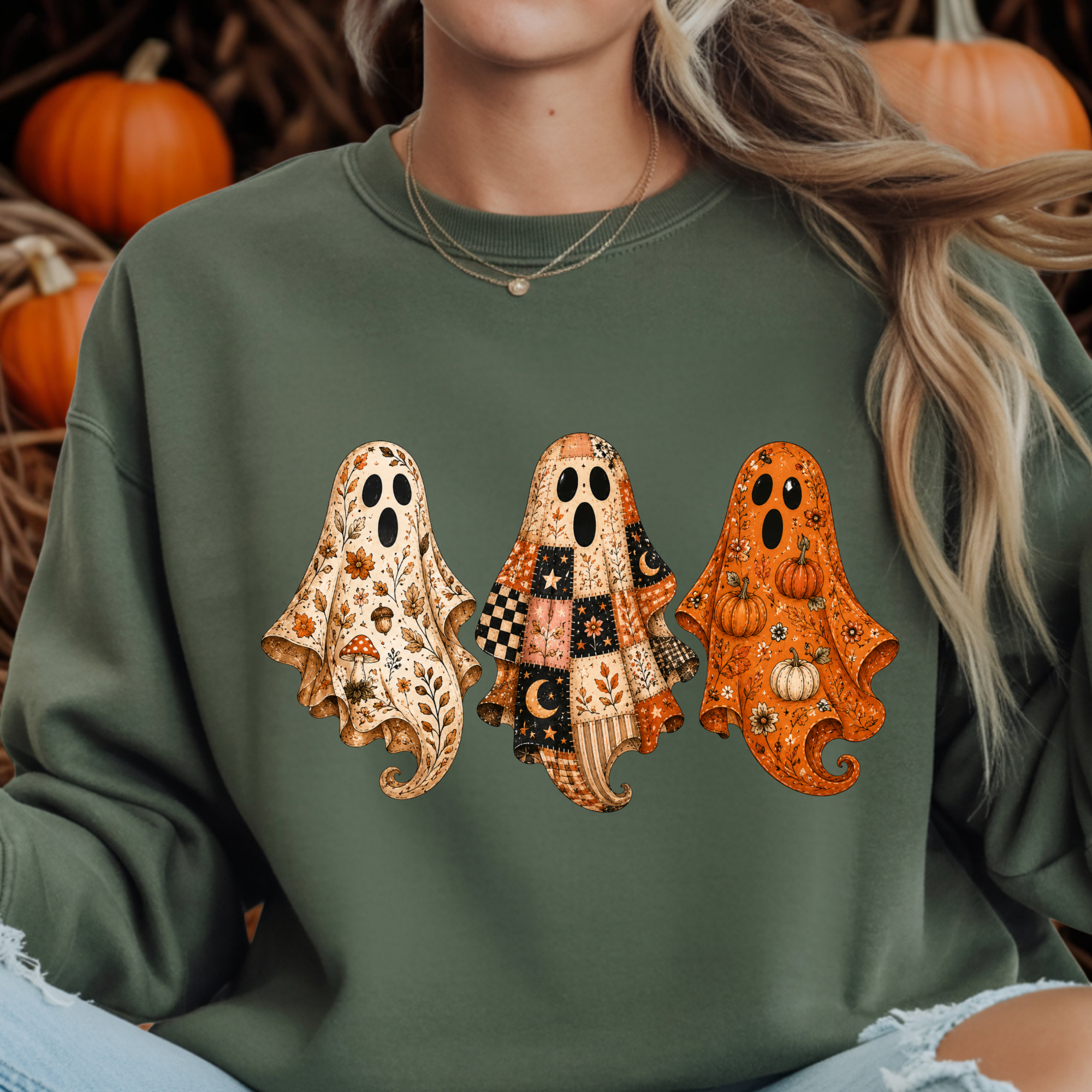 A person wearing an olive green sweatshirt with a graphic of three patterned ghosts in autumn colors, with pumpkins visible in the background.