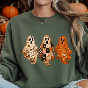 A person wearing an olive green sweatshirt with a graphic of three patterned ghosts in autumn colors, with pumpkins visible in the background.