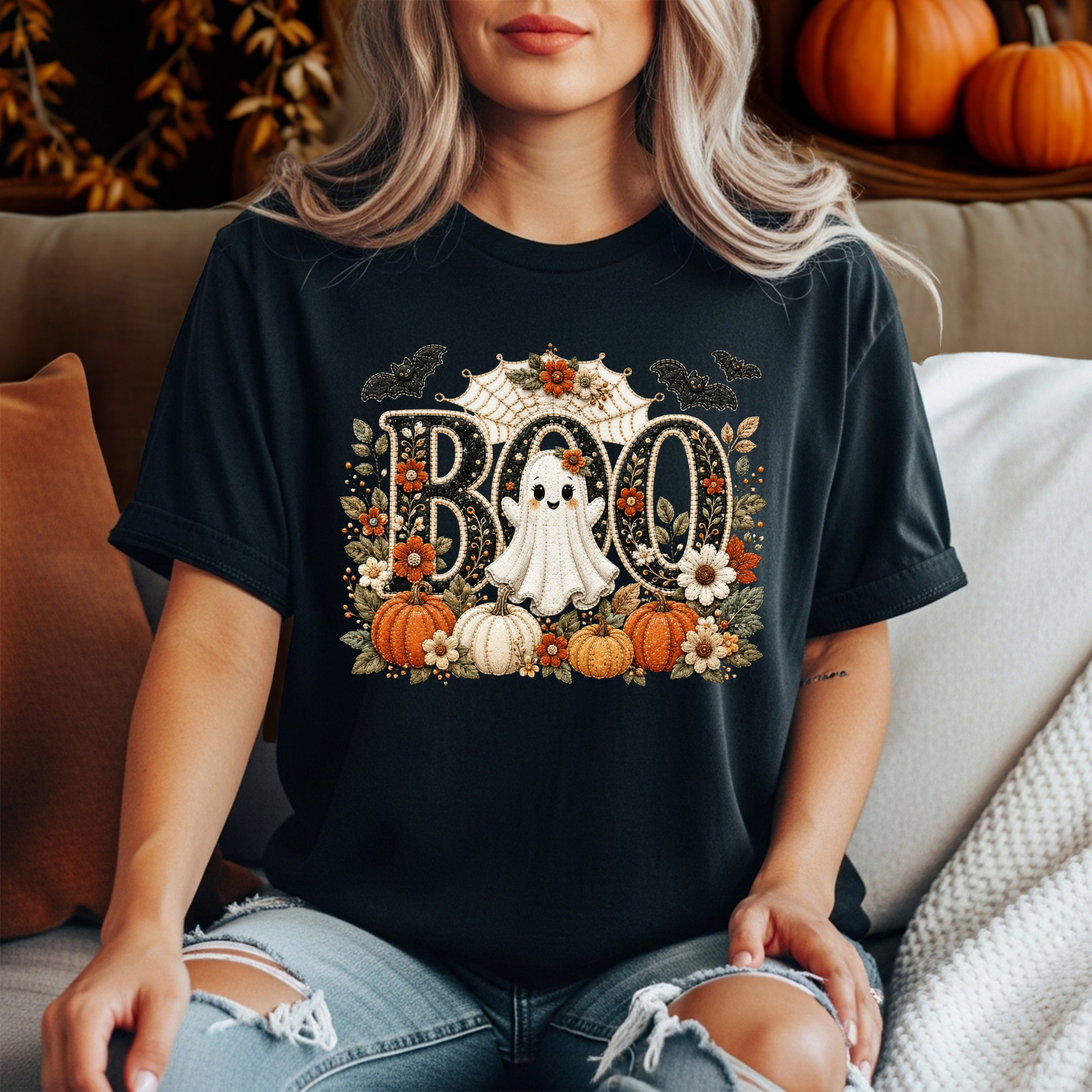 A woman wearing a black graphic t-shirt with a Halloween 'BOO' design featuring pumpkins, a ghost, and flowers, sitting on a couch.
