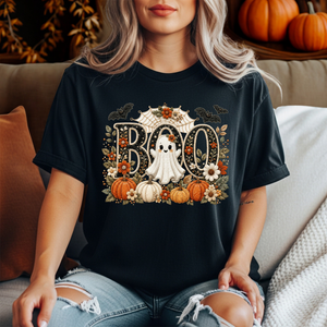 A woman wearing a black graphic t-shirt with a Halloween 'BOO' design featuring pumpkins, a ghost, and flowers, sitting on a couch.