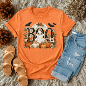 An orange short-sleeve graphic T-shirt laid flat on a shaggy faux-fur surface, styled with light blue jeans, tan sandals, and dried foliage. The shirt graphic reads BOO in large black letters with a white ghost, pumpkins, flowers, bats, and autumn leaves around it.