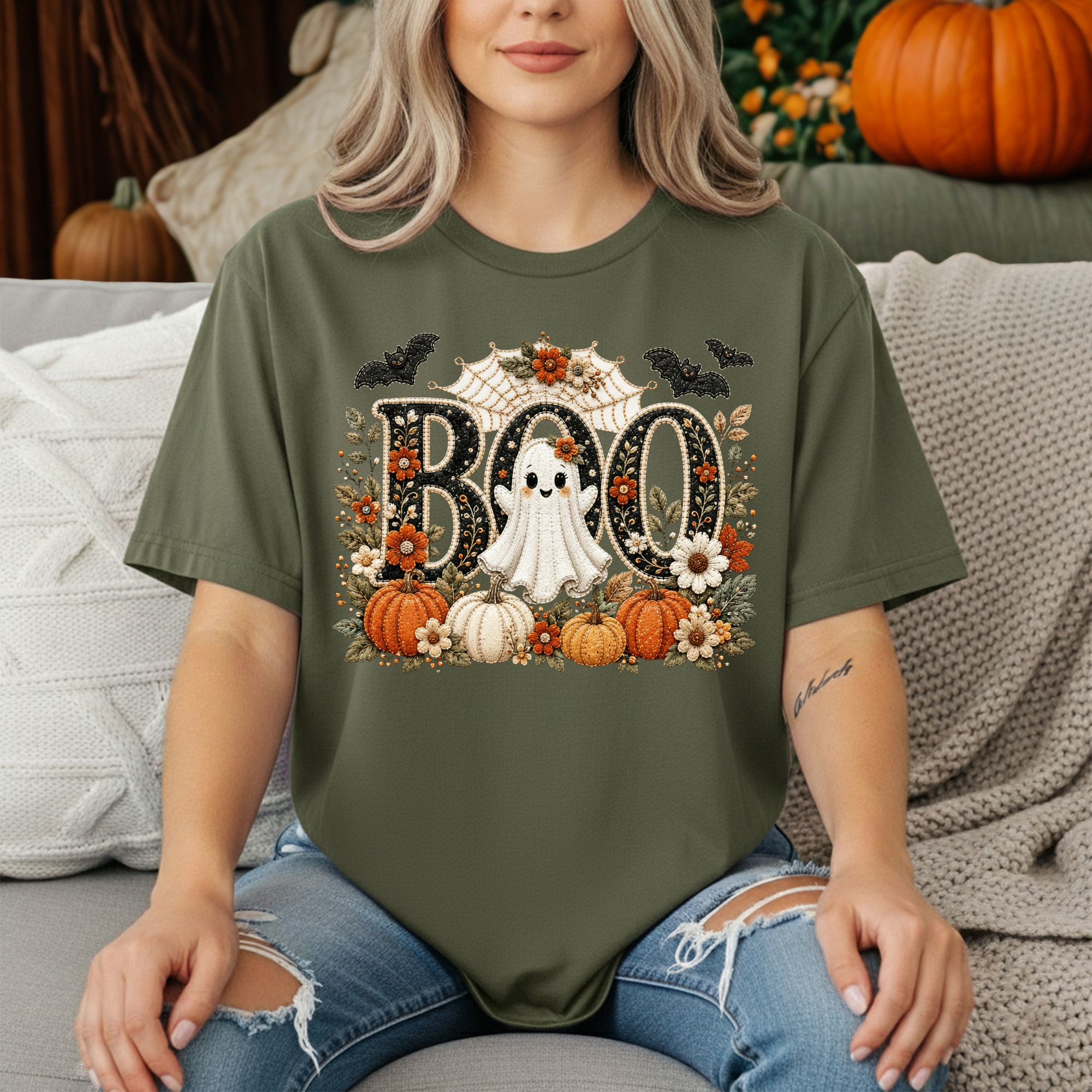 A woman wearing an olive green graphic t-shirt with a Halloween 'BOO' design featuring pumpkins, a ghost, bats, and flowers, sitting on a couch with pillows and pumpkins in the background.