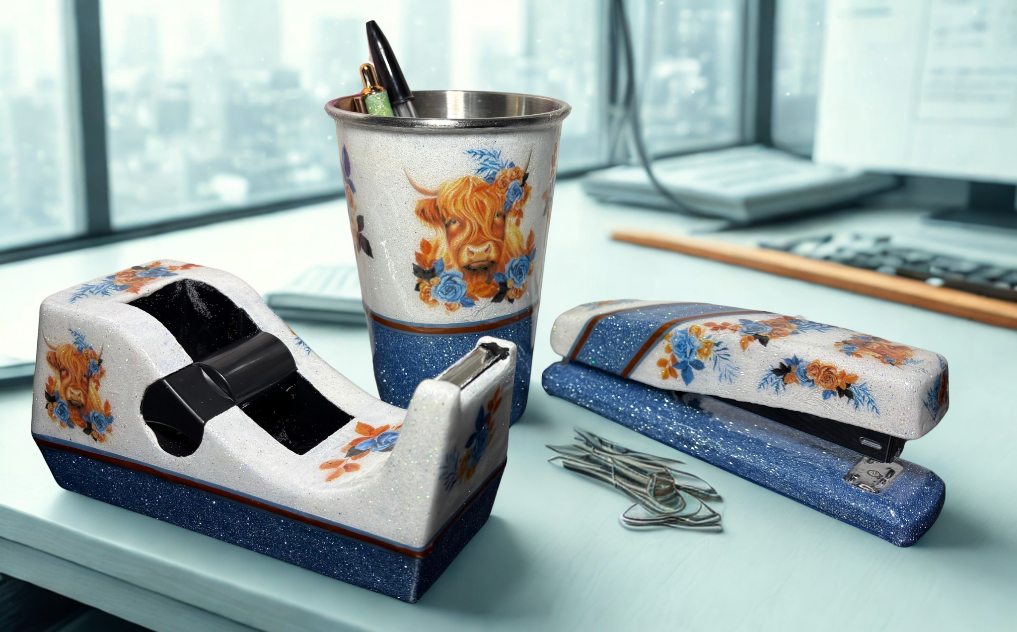 A set of decorative desk accessories including a tape dispenser, a cup holder, and a stapler, all featuring a white and blue speckled base with orange and blue floral designs and a Highland cow illustration. They are arranged on a light blue desk in front of a blurred office background.