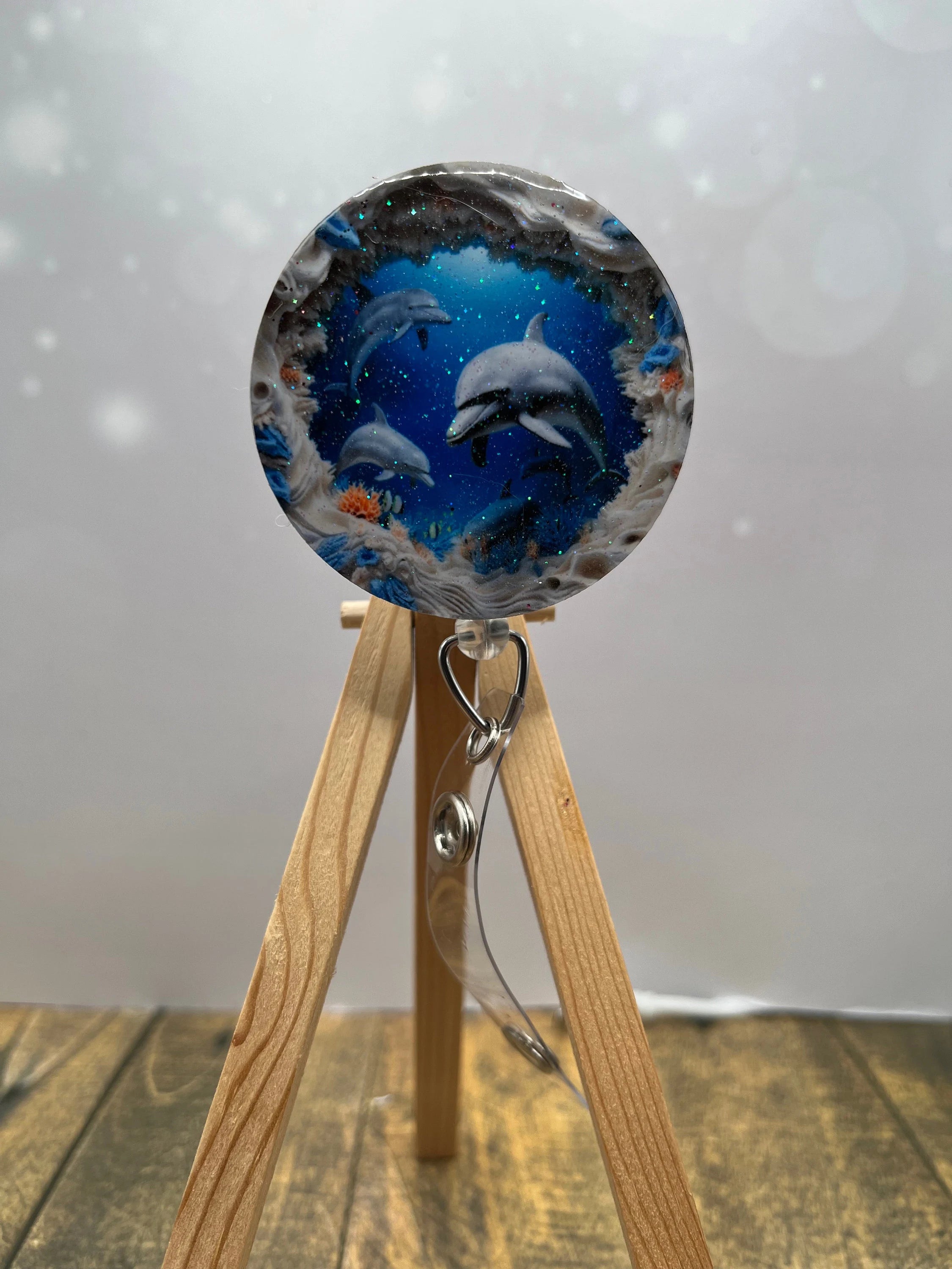 Dolphin badge pull with glitter featuring playful dolphins in a blue ocean scene on a wooden stand.