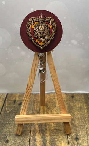 Gryffindor badge pull with glitter displayed on a wooden easel, featuring a lion crest design.